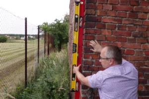 Ekspert budowlany dokonuje pomiaru narożnika budynku przy użyciu poziomic elektronicznych, oceniając deformacje konstrukcji na terenie oddziaływań szkód górniczych.